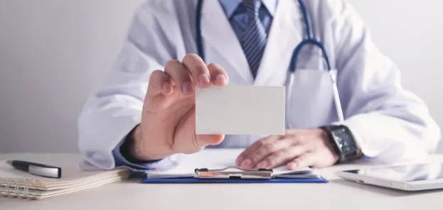 A doctor holds up a card while sitting at a desk with a clipboard and notepad